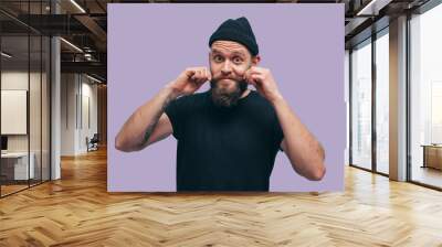 Handsome hipster guy with beard smiling happily, dressed casually with black beanie on head isolated over a blue studio background. The concept of people, crazy emotions. Wall mural