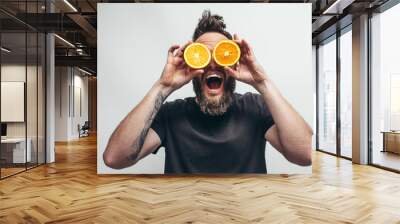 Young bearded hipster man holding slices of orange in front of his eyes. Crazy emotions Wall mural