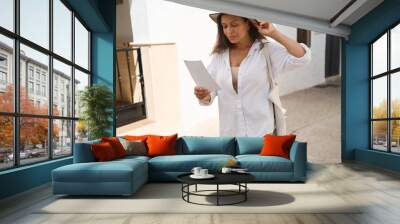 Woman in casual attire reading a document while strolling down a sunny cobblestone street, wearing a stylish hat, creating a blend of leisure and curiosity in a serene urban setting. Wall mural