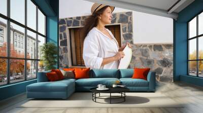 Woman in white blouse and straw hat enjoying a sunny day outdoors, standing by a stone wall, holding a hand fan with relaxed and content expression. Wall mural