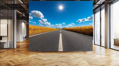 A straight asphalt road cutting through a golden wheat field under a bright blue summer sky. Wall mural