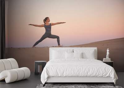 Beautiful young woman practices yoga asana Virabhadrasana 2 - warrior pose 2 in the desert at sunset Wall mural