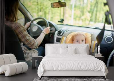 Young beautiful woman driving a car. On a front seat mounted child safety seat with a pretty 1 year old toddler boy. Child transportation safety. Wall mural