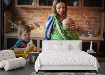 Young family, mother with two children, preschool boy and baby in sling cooking together in a sunny kitchen. Wall mural