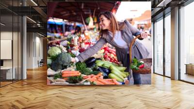 Happy charming Caucasian brunette holding basket and buying vegetables at farmer's market. Wall mural