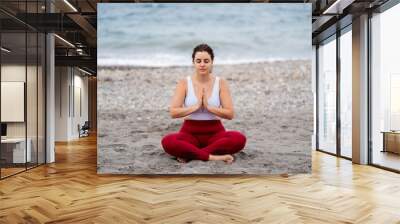 Beautiful lady in lotus pose doing yoga at the beach with palms together and closed eyes Wall mural