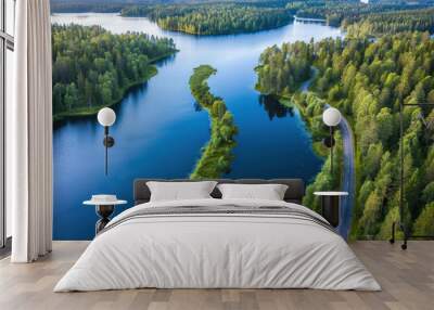 Bird's-eye view of a tranquil road meandering through vibrant green forests and alongside shimmering blue lakes in Finland, bathed in summer sunlight. Wall mural