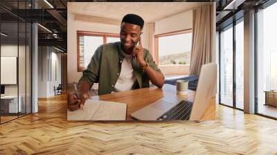 Young adult black African American male smiling while on a phone call. Writing in his notepad, working from laptop at home in modern apartment. Wall mural