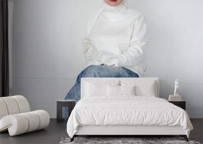 A Muslim woman in a white hijab and a white blouse, in a denim skirt, sits in a high chair. Stylish Muslim. Copy space. Wall mural