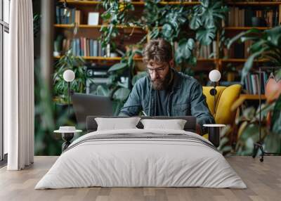 Focused bearded man working on a laptop in a cozy home office surrounded by plants, concept of remote work and creative workspace Wall mural