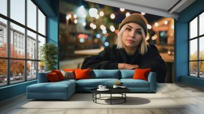 A woman wearing a brown hat and a black sweater is sitting at a table. She is looking at the camera with a serious expression. The scene takes place at night, with lights illuminating the area Wall mural