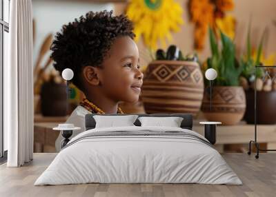 A young child is sitting in front of a table with a vase and a potted plant. The child is wearing a necklace and a white shirt Wall mural