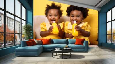 Two Nigerian baby girls, aged 2 and 1, sitting on a yellow sofa, drinking milk from baby feeding bottle. Wall mural