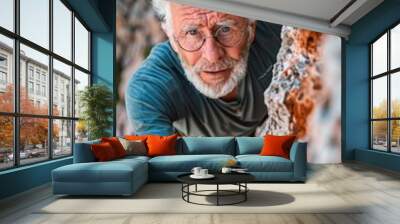 Mature white haired man climbing up a cliff wearing glasses Wall mural