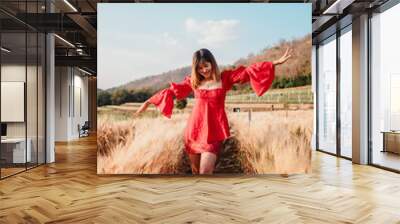 A cheerful woman in a red dress stands in a wheat field, enjoying the sunny day with a blue sky and hills in the background. Wall mural