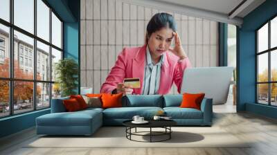 A woman in a pink blazer looks stressed while holding a credit card and staring at her laptop in an office environment. Wall mural