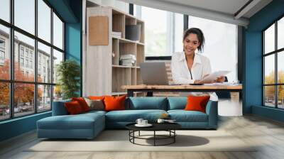 A young professional woman in a modern office setting, working at a desk with a laptop and clipboard, smiling confidently. Wall mural