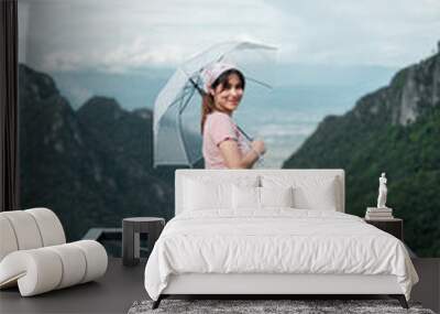 A young woman stands on a balcony with an umbrella, overlooking lush green mountains under a cloudy sky. Wall mural