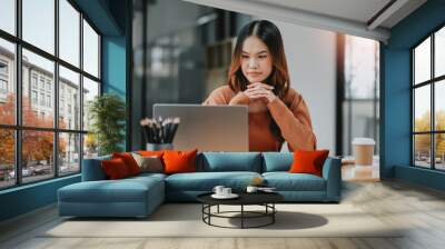 Contemplative young woman deeply focused while working on her laptop at a well-organized office desk. Wall mural