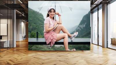 Smiling woman in casual attire holding a clear umbrella, seated on a bench with lush green mountains in the background. Wall mural