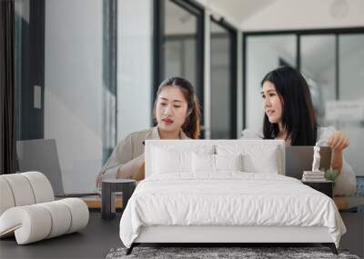 Two professional women engaged in discussion, with one writing in a notebook and the other using a laptop at an office table. Wall mural