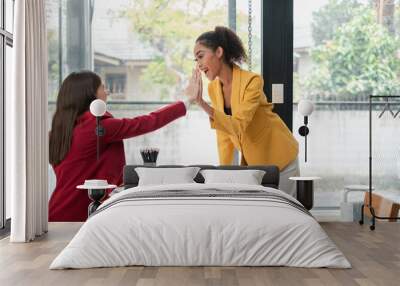 Two professional women in vibrant blazers high-fiving at a desk, celebrating success in a contemporary office environment. Wall mural