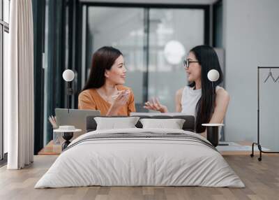 Two women smiling and talking at a desk in a modern office setting, with a laptop and paperwork. Wall mural