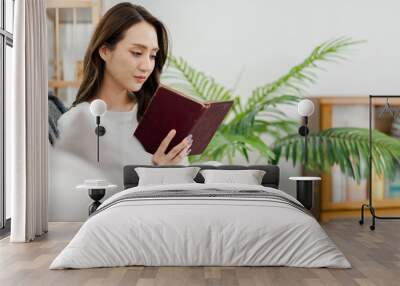 young woman reading a book comfortably on a couch in a well-lit room with houseplant decorations. Wall mural