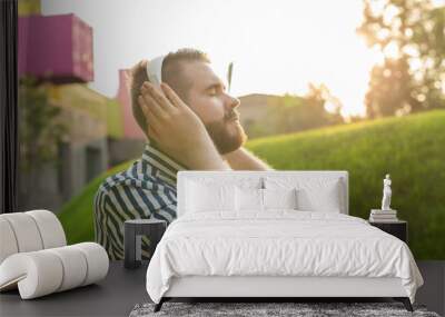 Handsome relaxed young hipster man with a mustache and beard is listening to favorite music at the earphones while walking through the park. Music subscription concept and online radio. Wall mural