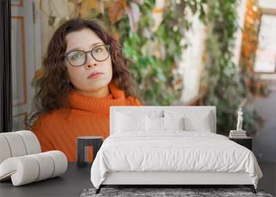 Portrait of carefree pensive young woman smiling with urban background. Cheerful latin girl wearing eyeglasses in the city. Happy brunette woman with curly hair and spectacles smiling. Copy space and Wall mural