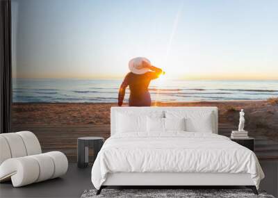 Travel, holiday and people concept - woman tourist sitting near the sea on the red suitcase and in hat and watching sunset Wall mural