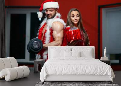 Christmas. Handsome muscular man in Santa Claus suit with dumbbells and fitness woman in red suit with gift . GYM. Wall mural