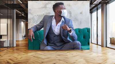 COVID-19. Confident young African man is wearing a protective medical face mask to prevent infection with coronavirus and pandemic sitting on a green sofa in a grey suit against a concrete wall Wall mural