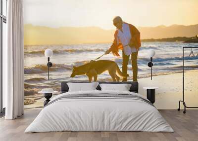 Happy attractive senior woman with her german shepard dog walking on the beach at autumn sunset Wall mural