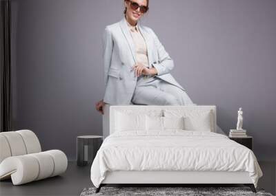 Fashion photo of a beautiful elegant young woman in a pretty soft blue suit, jacket, pants, trousers, white boots, stylish sunglasses on gray background. Studio shot, portrait. Model sits on cube Wall mural