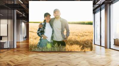 A couple of farmers in plaid shirts and caps stand embracing on agricultural field of wheat at sunset Wall mural