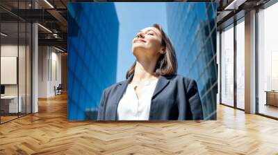 A confident businesswoman stands with closed eyes, basking in the sun amidst towering city buildings, reflecting a sense of peace and accomplishment. Wall mural