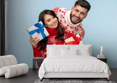 Beautiful couple dressed in Christmas sweaters holding gift boxes smiling over blue background. Wall mural