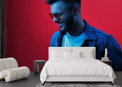 Cheerful stylish hipster bearded young man wearing yellow sunglasses and blue jeans jacket posing in neon light over red background. Wall mural