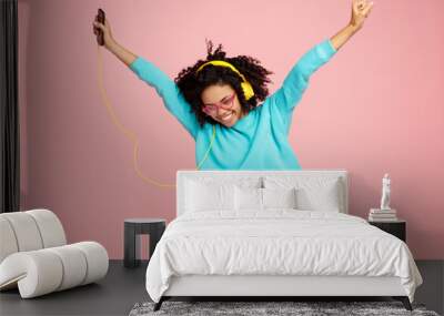 Pretty african american young woman with bright smile dressed in casual clothes, glasses and headphones dance over pink background. Wall mural