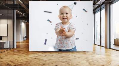 A little cute happy girl rejoices and laughs on her first birthday on a white background. Slapstick with confetti, a balloon filled with helium, a silver cap. Wall mural