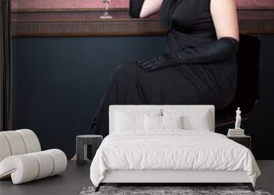 leggy woman wearing long black dress and long black gloves is sitting on a bar chair with glass of cocktail and smoking from cigar-holder Wall mural