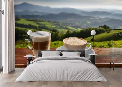 a cup of cappuccino and a glass of latte macchiato on a wooden table overlooking a hilly landscape Wall mural