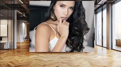 young gorgeous asian filipino woman with tan smooth skin and long curly hair wearing a lingerie set with a white lacy bra while posing with her finger in her mouth on a wooden bed with white sheets Wall mural