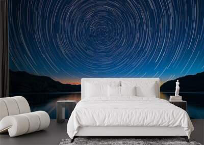 long-exposure shot of star trails swirling above a calm lake and mountainous horizon at twilight, reflecting the cosmic dance of the stars in the water below. Wall mural