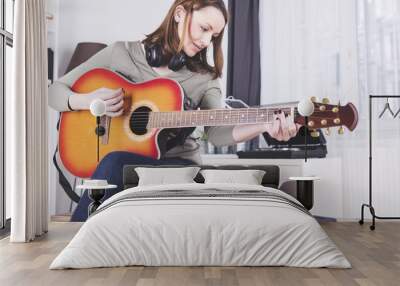 Concentrated brown haired girl with headphones around her neck playing some records on guitar while using a tablet Wall mural