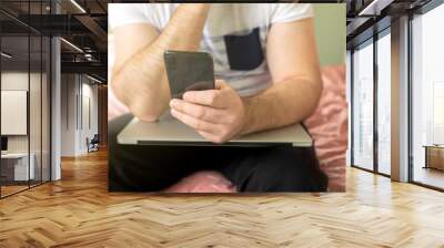 Adult male or man working from home with his laptop on his knee and with a phone in his hands reading something. One hand on his chin. Wall mural