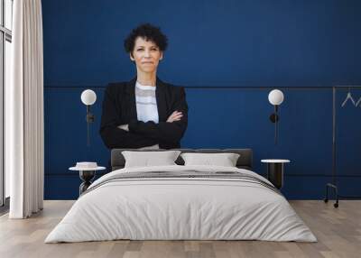 Portrait of confident mature female entrepreneur standing with arms crossed against blue wall Wall mural