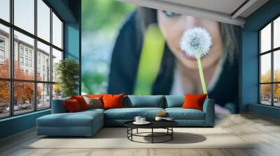 Close up portrait of a young woman with a dandelion laying on the grass. Positive girl having fun outside in a sunny day. Focus on the flower. Wall mural