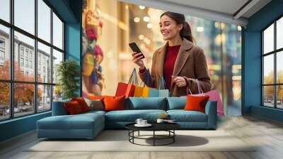 A smiling woman in a brown coat holds colorful shopping bags and checks her smartphone while walking on a city street. Wall mural